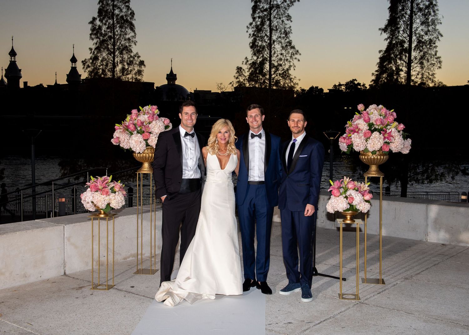 Bride and groom pose with two guests at an outdoor wedding ceremony at dusk with pink floral arrangements.