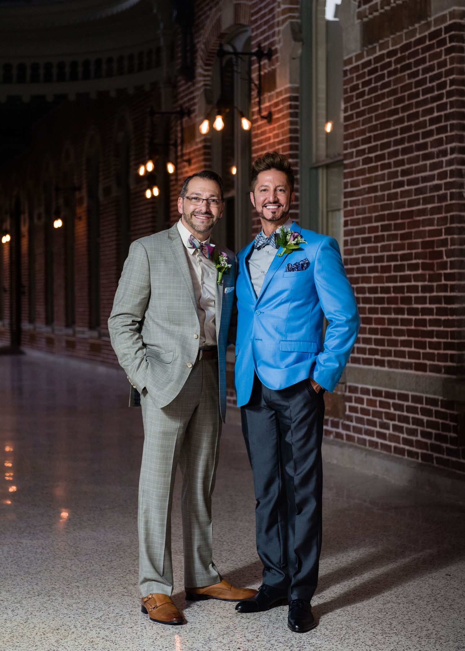 Two grooms in suits pose together in a brick hallway with warm lighting at their wedding celebration.