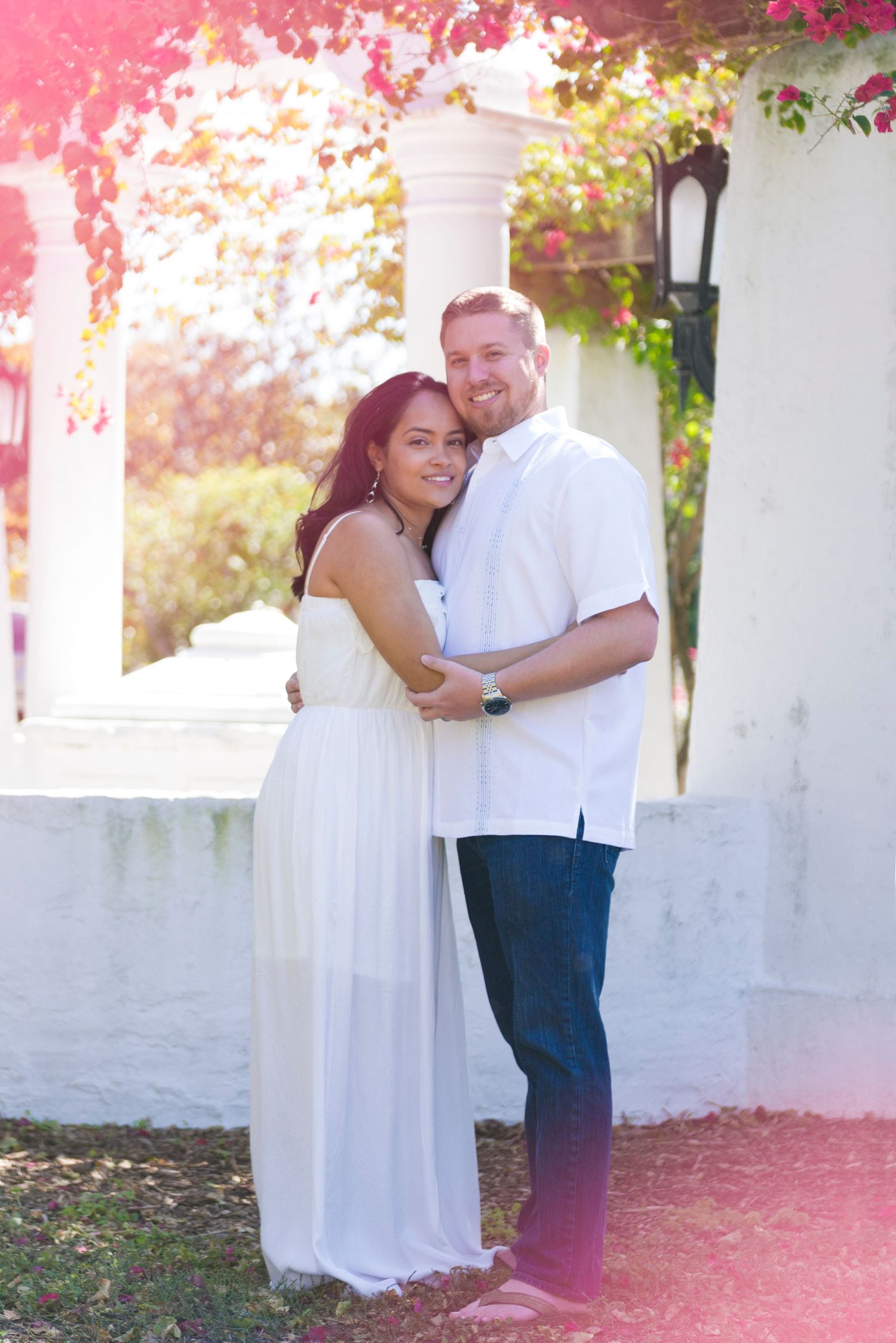 Couple embracing outdoors near white columns with pink bougainvillea, woman in white dress, man in white shirt and jeans.