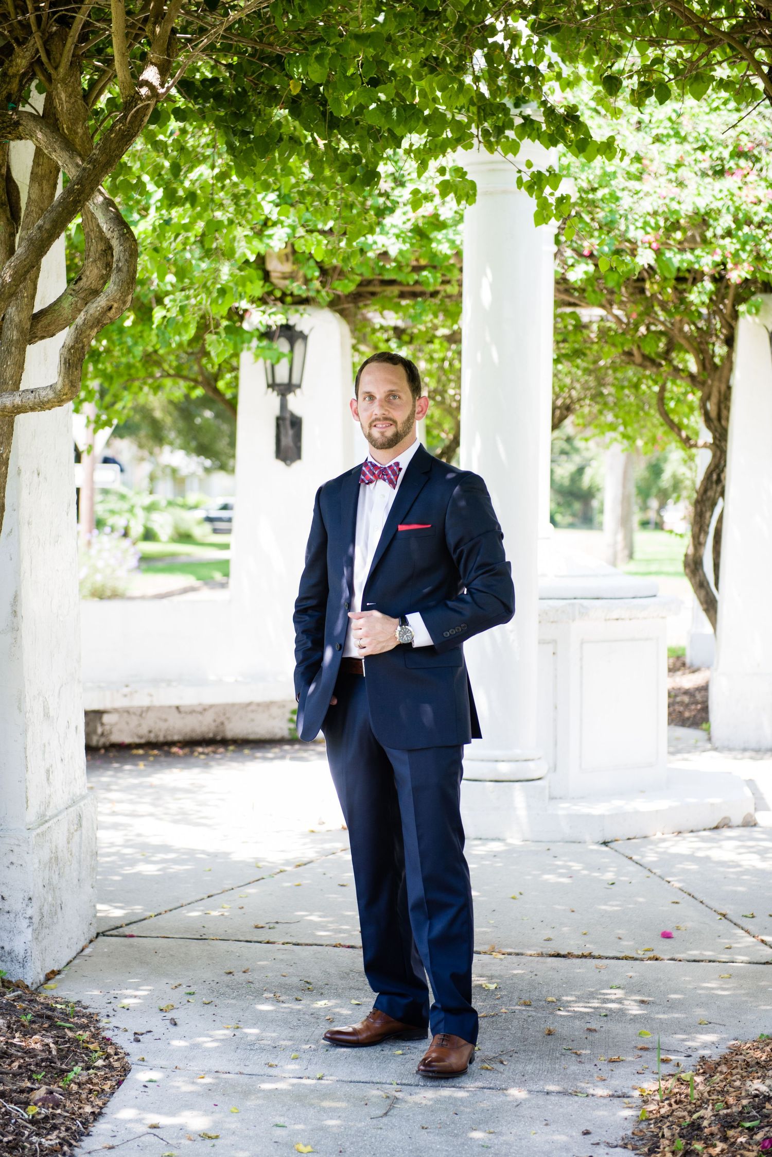 Handsome groom in navy blue suit with burgundy bow tie posing outdoors under lush green trees.