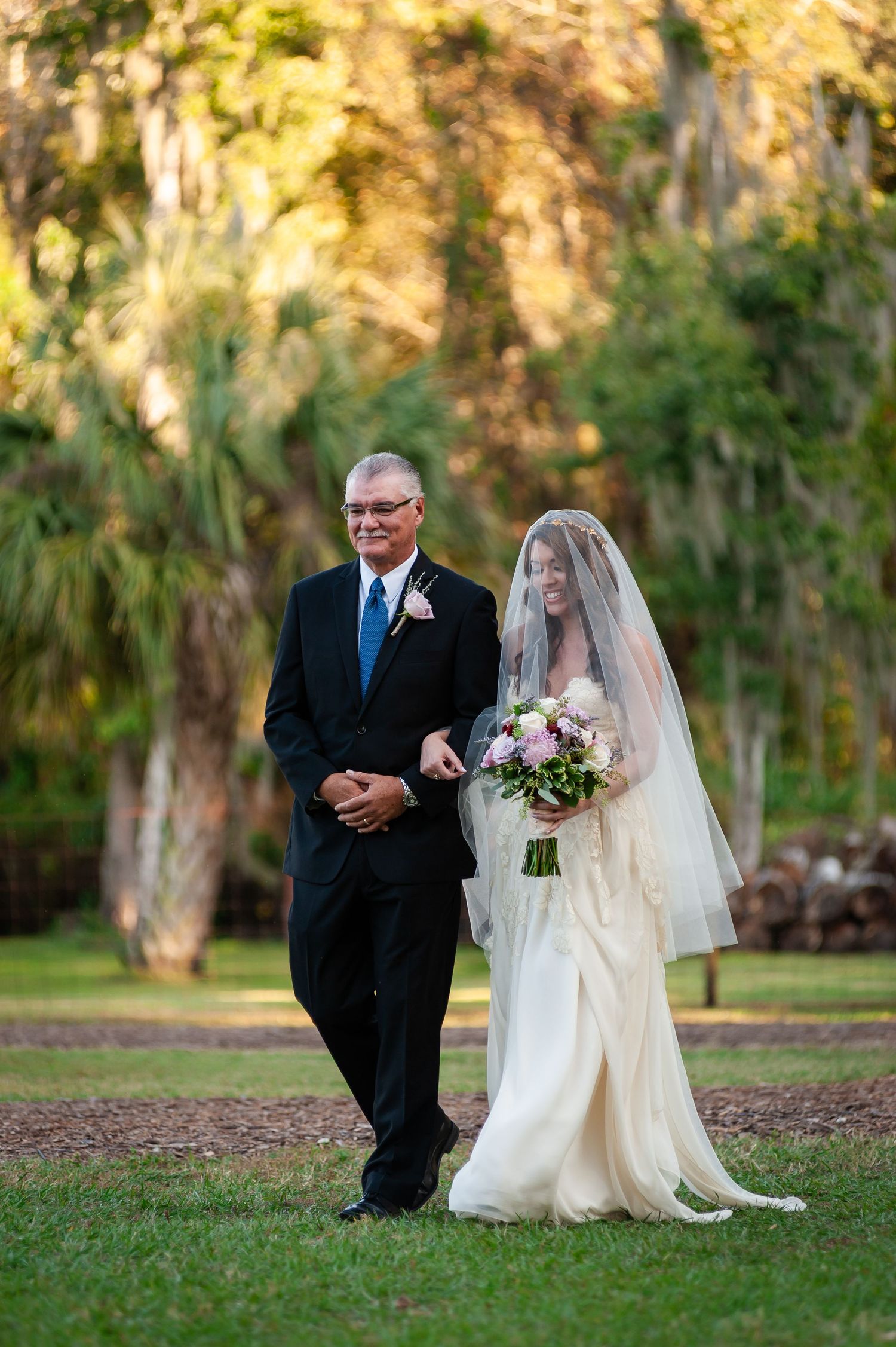 Bride in white gown and veil walks with suited man through lush green outdoor wedding ceremony venue.