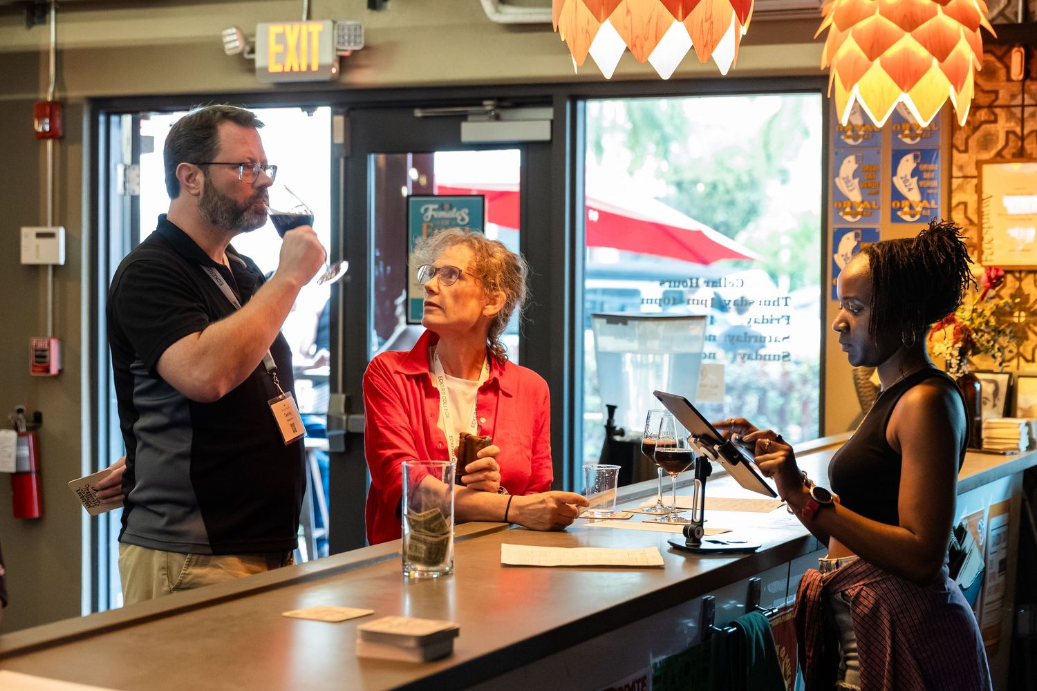 Three people conversing at a bar counter indoors, with one holding a drink and another a tablet.