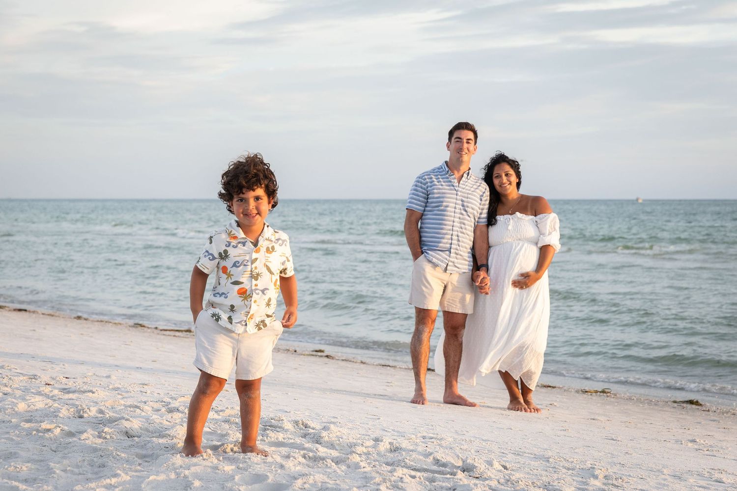 Family of three poses on a white sand beach, with a young boy in front and pregnant mother in a white dress.