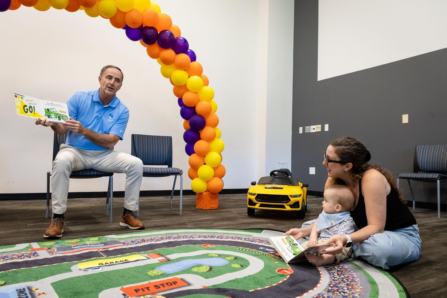 Man reads colorful book to woman and baby sitting on play rug under balloon arch in bright community room.