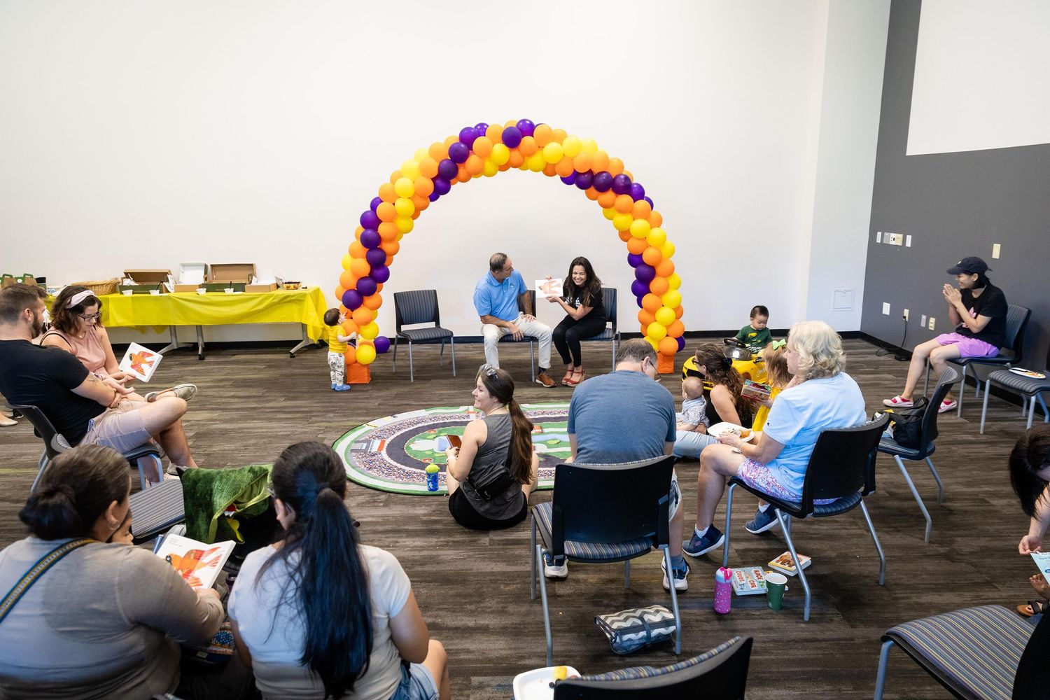 People seated in a circle under a purple and gold balloon arch during a community event in a modern venue.