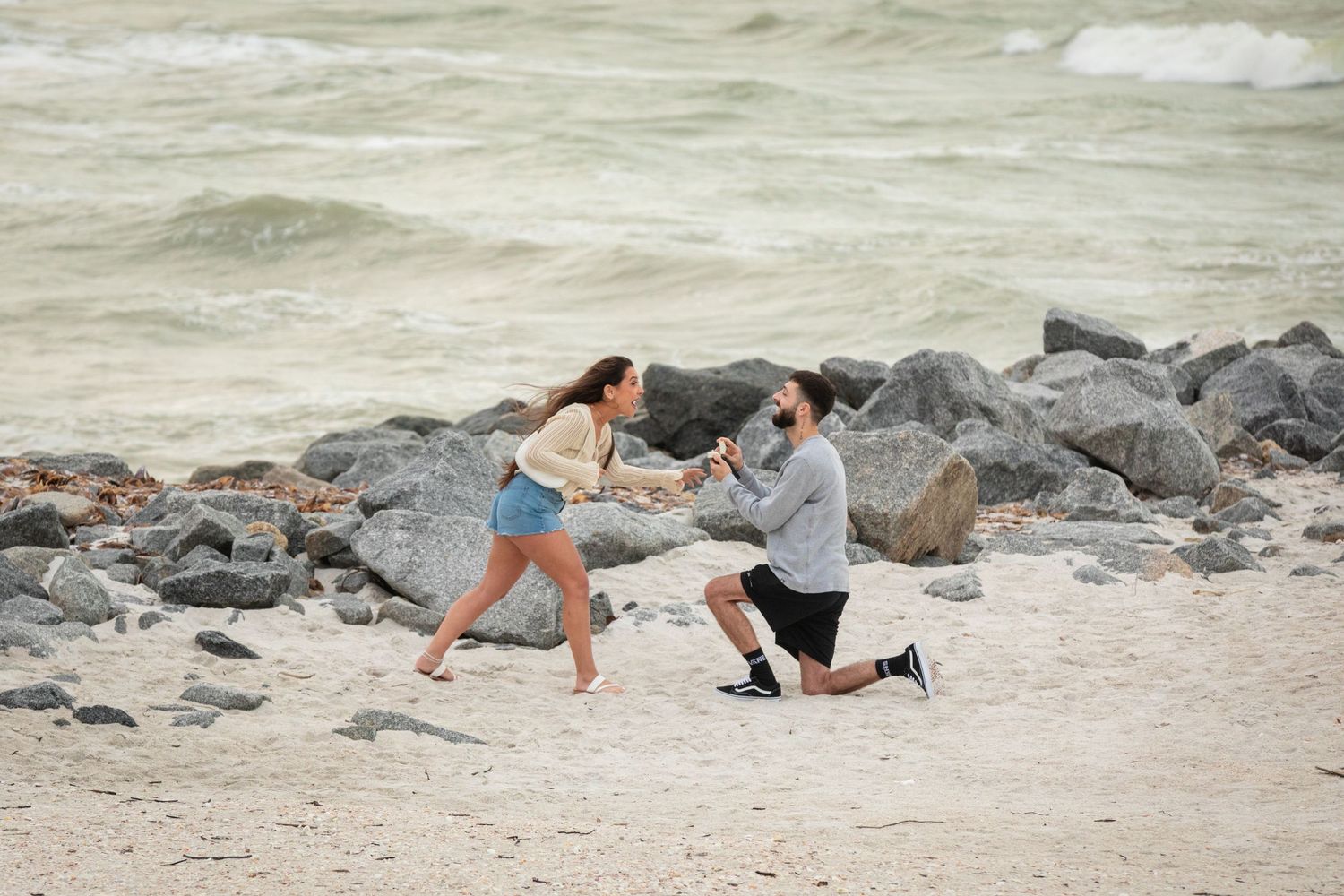 Man proposes on one knee to woman on sandy beach with rocky shoreline and ocean waves in background.