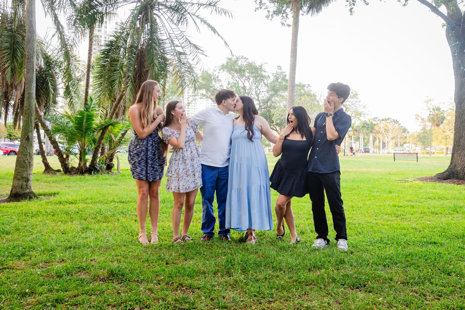 Group of six friends laughing and embracing outdoors on green grass surrounded by palm trees in a sunny park.