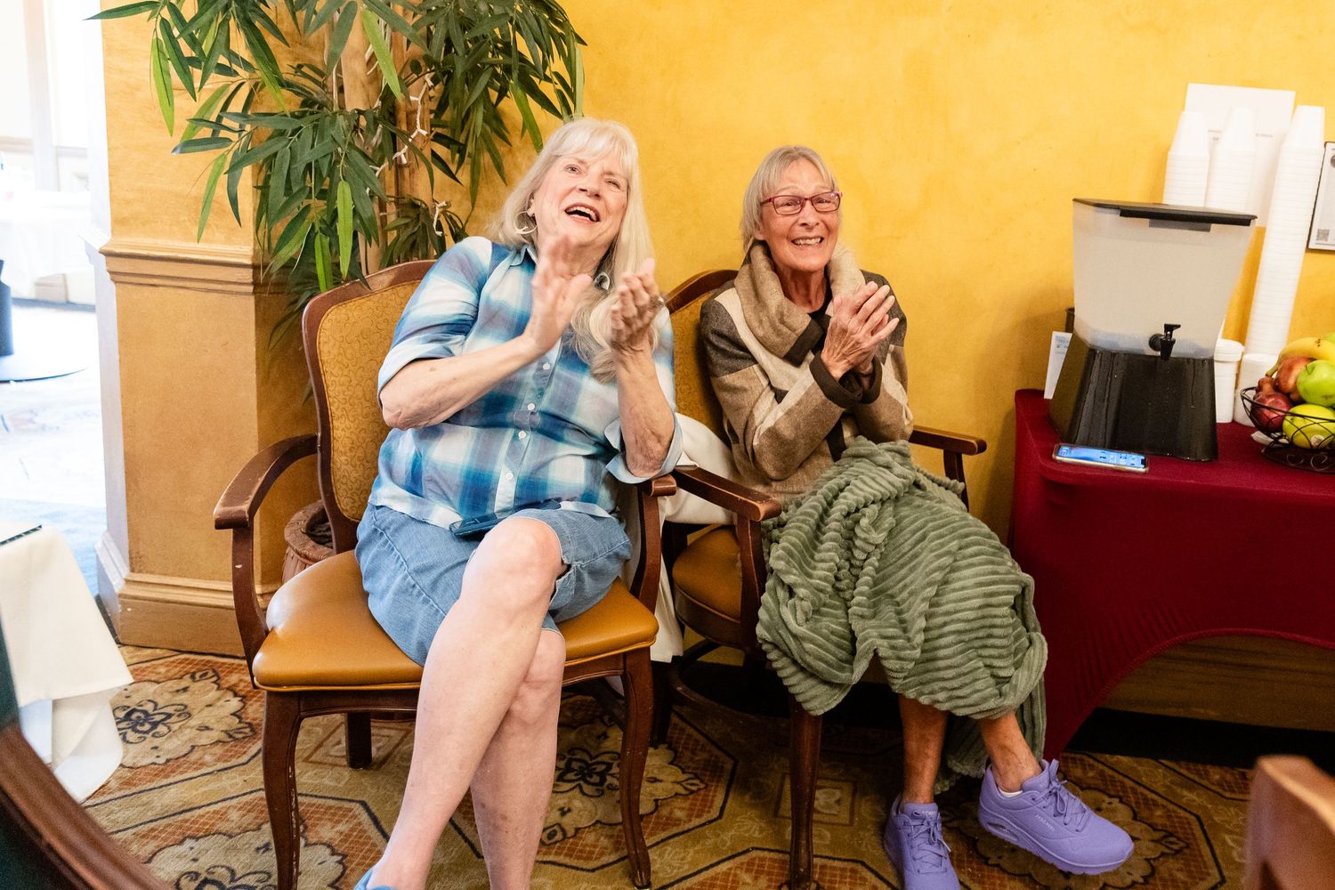 Two elderly women laugh and clap joyfully while seated in chairs at a Pride community celebration event.