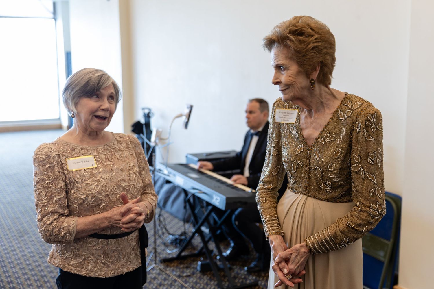 Two elegantly dressed women share a laugh at a formal event while a pianist performs in the background.