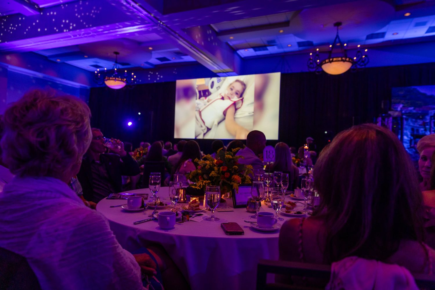 Guests seated at elegant round tables watch a presentation on a large screen at a formal gala dinner event.