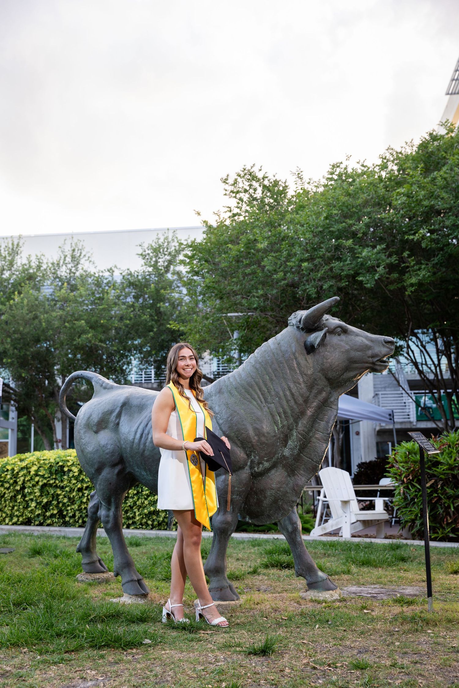 Graduate in white dress and yellow sash poses beside a large bronze bull statue outdoors.