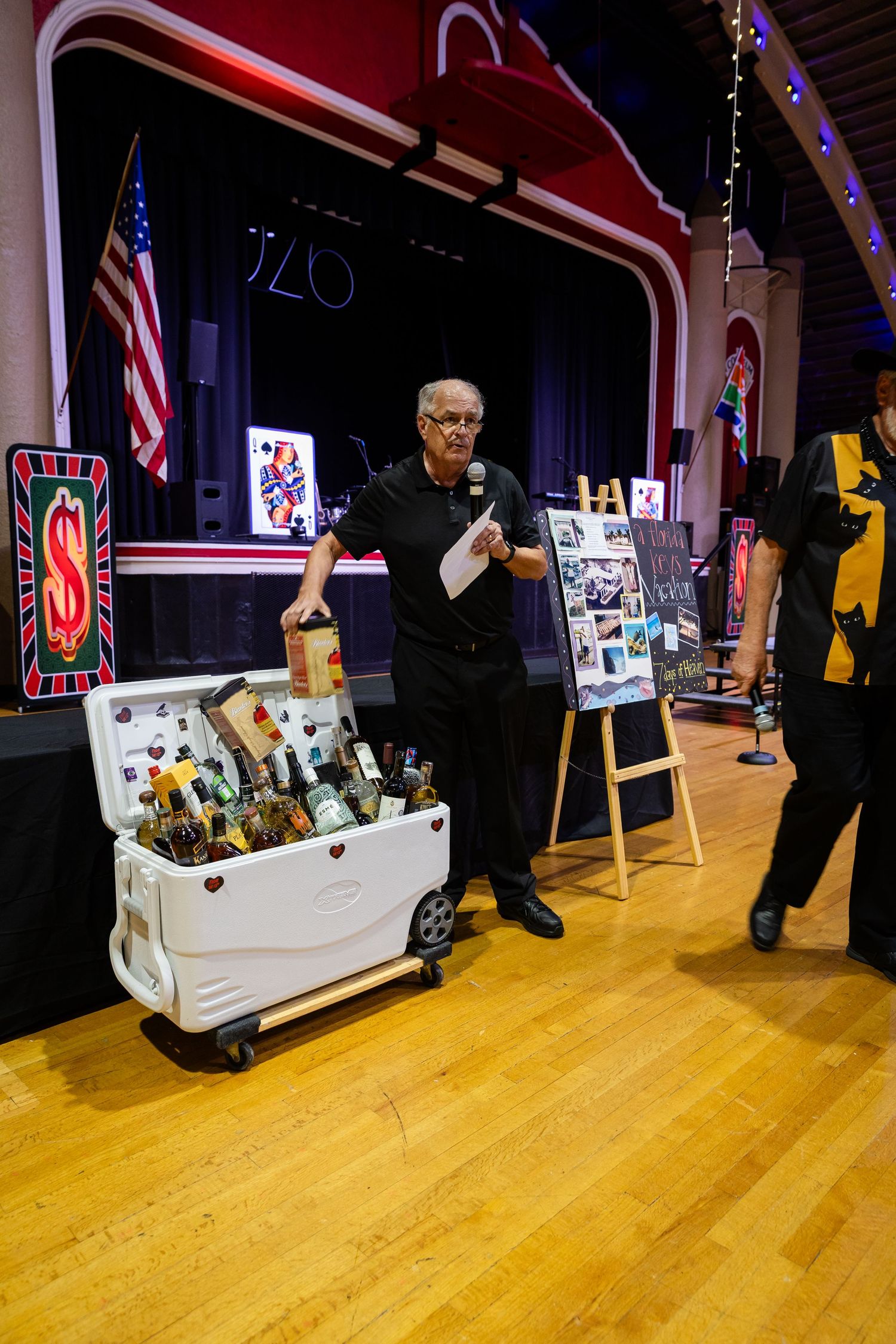 Man in black holding microphone stands beside a cooler filled with bottles at an indoor event with stage backdrop.