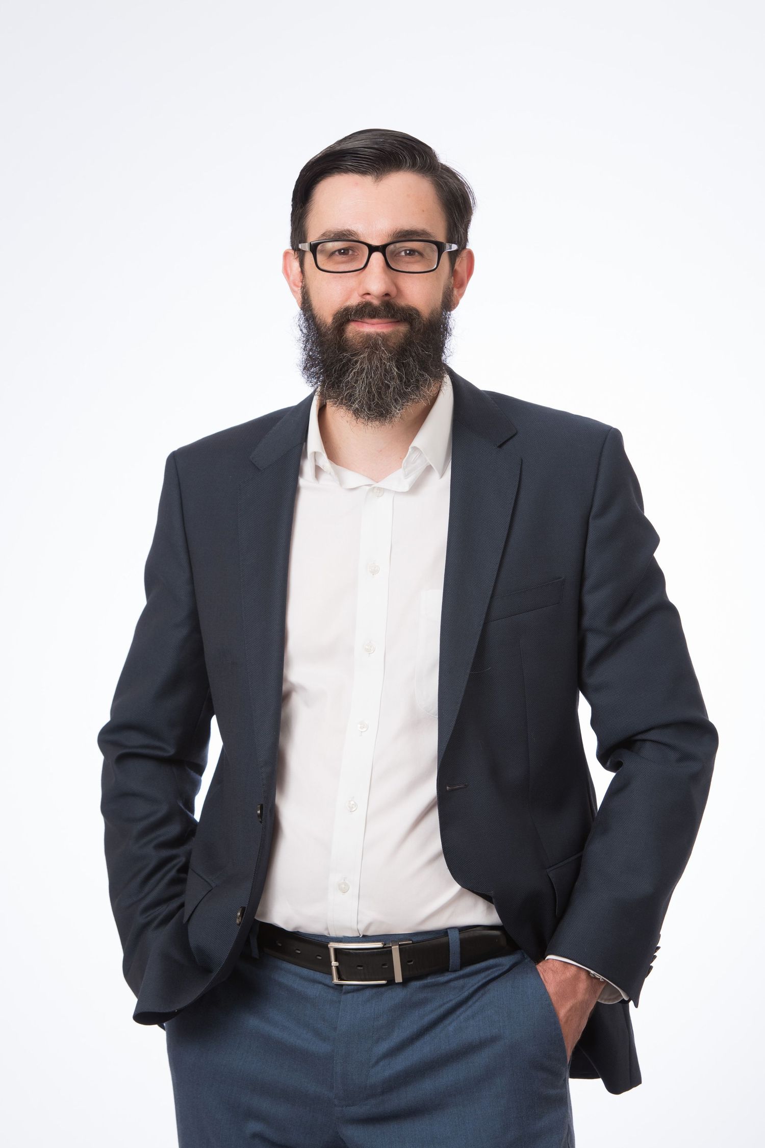 Bearded businessman in dark blazer and glasses poses confidently with hands in pockets against white background.