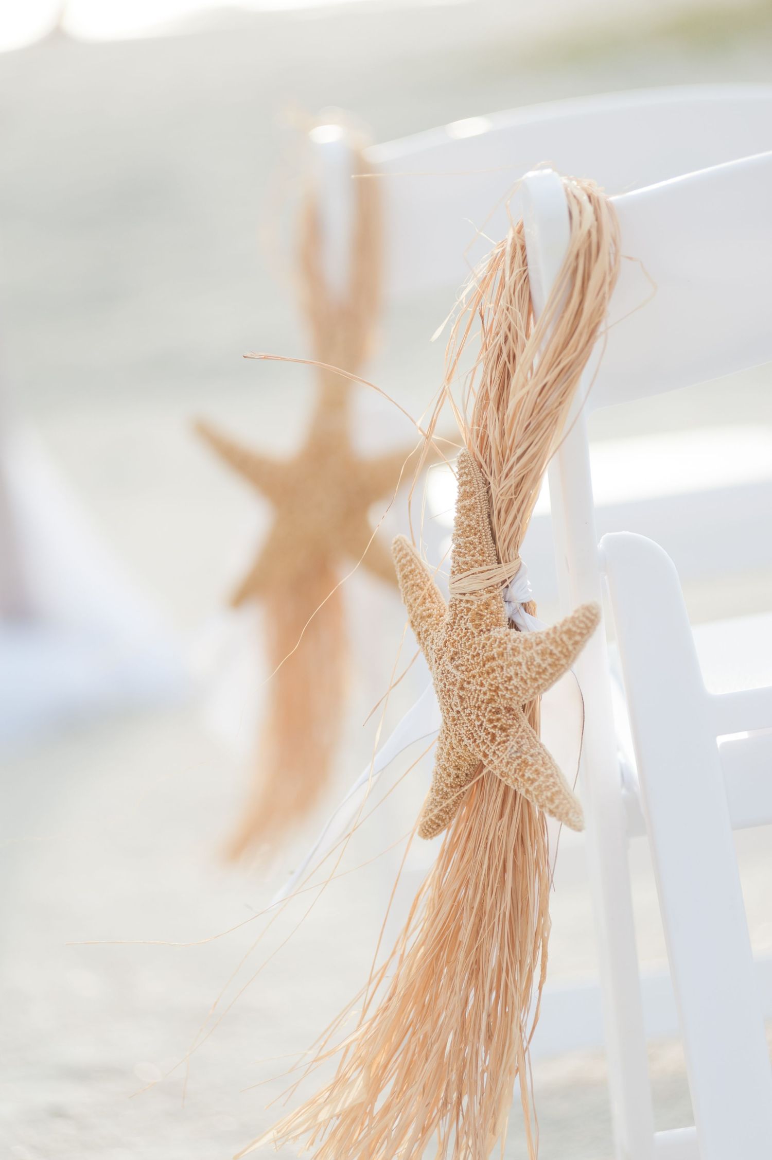 Starfish tied with natural raffia ribbon decorating white chairs at a beach wedding ceremony.