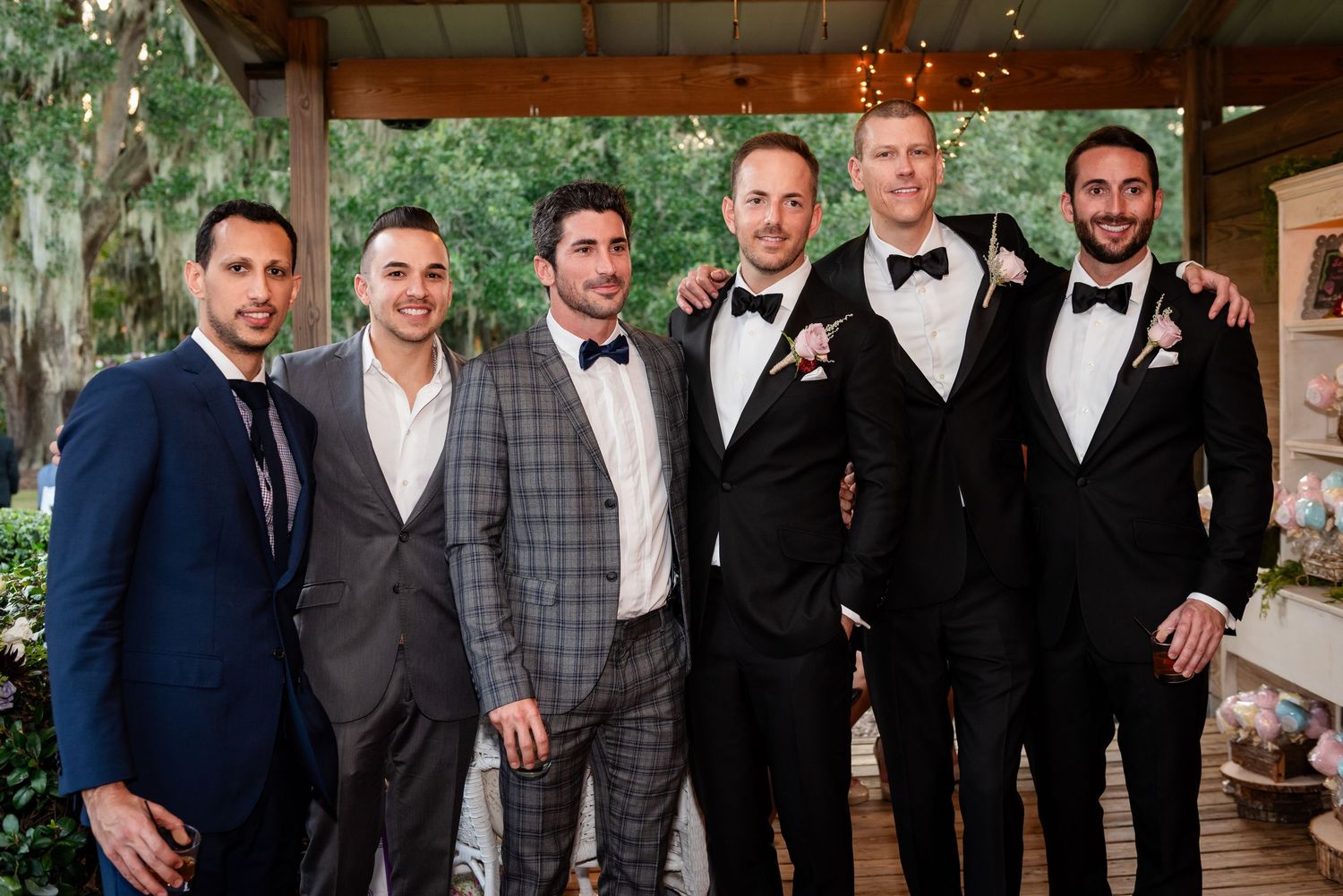 Six male wedding guests in suits and tuxedos posing together under a covered outdoor venue patio.