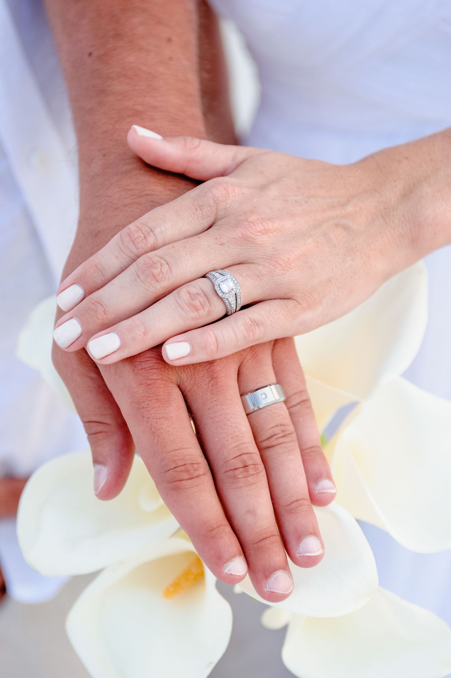 Two hands with wedding rings resting on white orchids, showcasing a diamond engagement ring and a silver band.