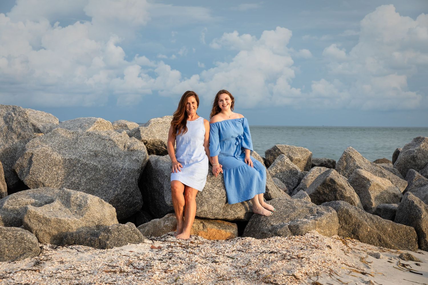 Two women in blue dresses posing on rocky shoreline with ocean and cloudy sky backdrop.