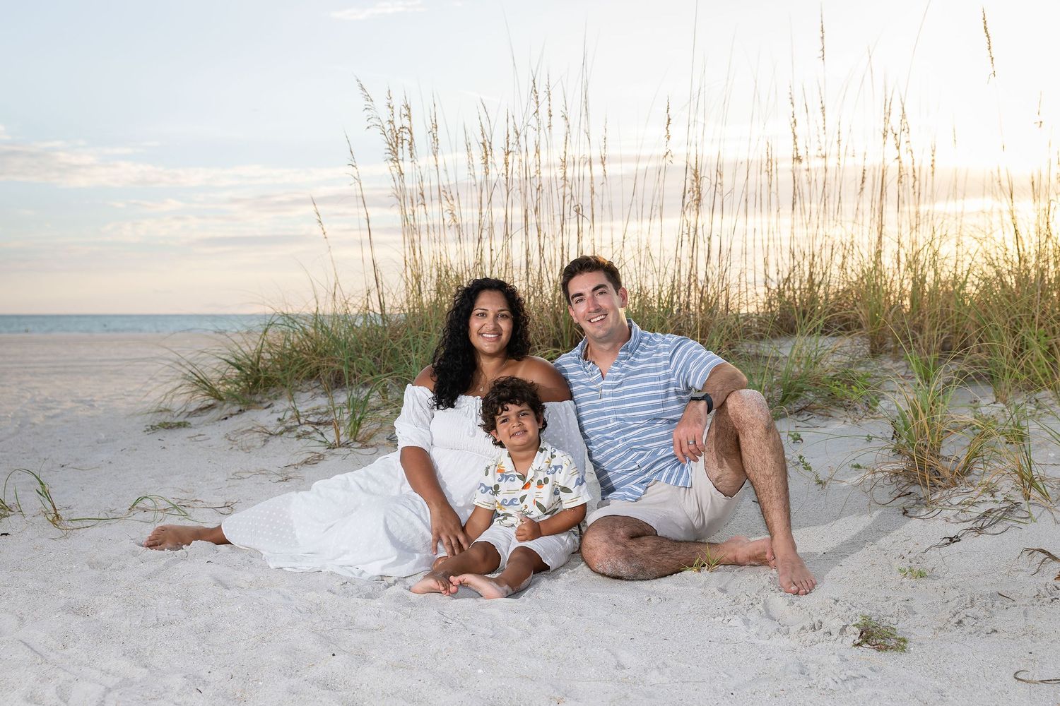Family of three smiling together on a sandy beach at sunset, sitting near sea oats on white sand.