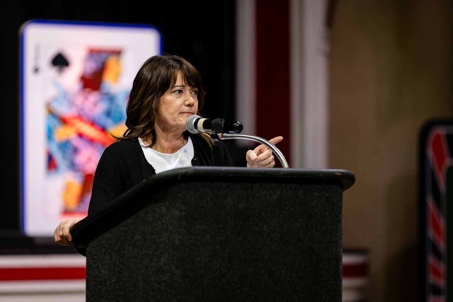 Woman speaking at podium with microphone, large playing card backdrop visible behind her at indoor event.