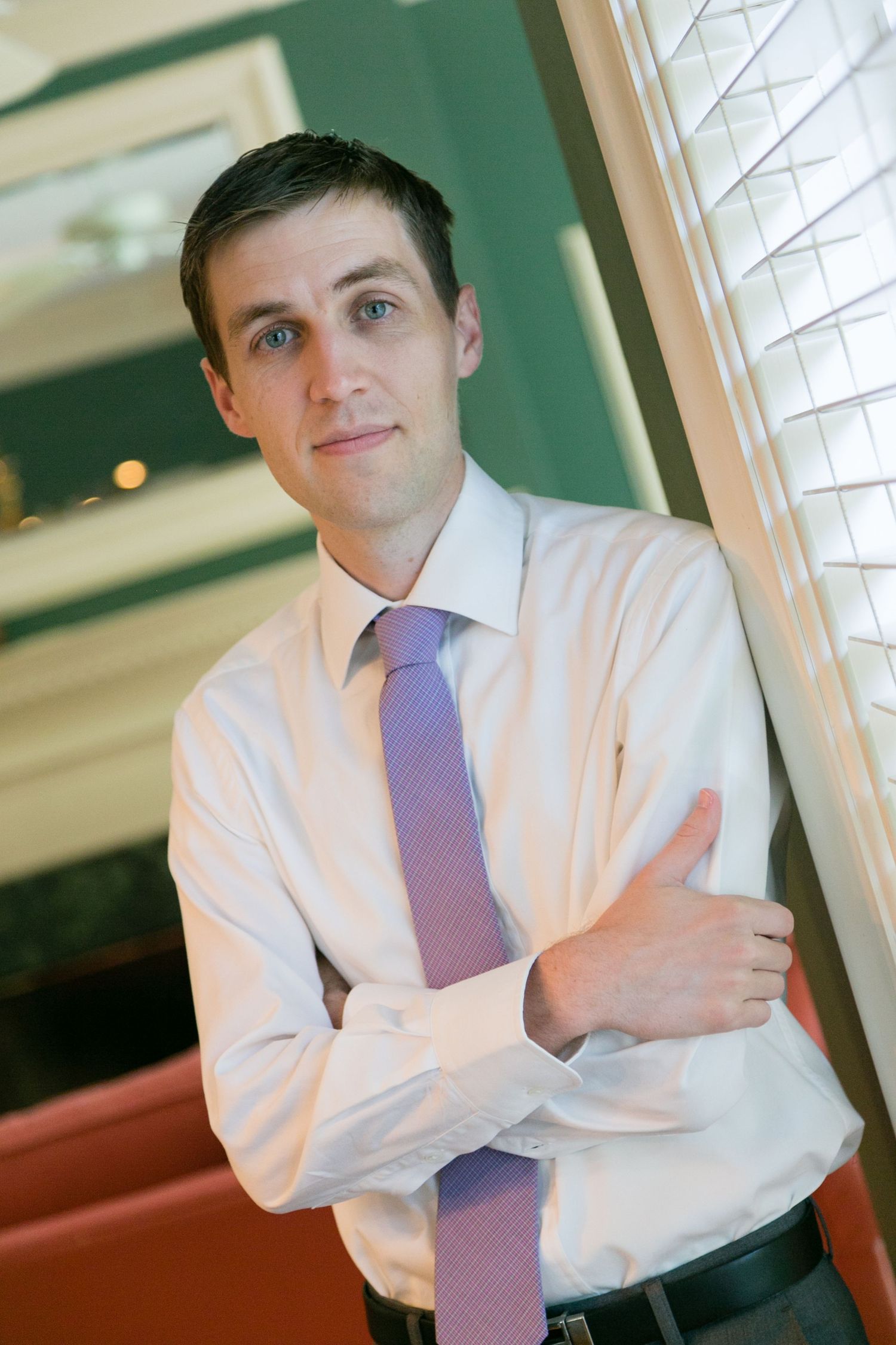 Handsome groom in white dress shirt and purple tie leaning against window before wedding ceremony.