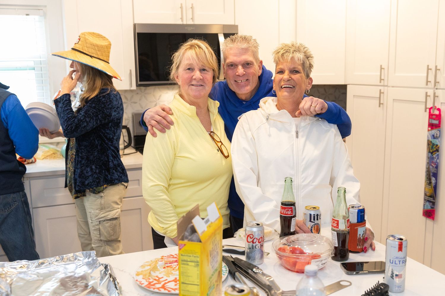 Three people smiling and posing together in a kitchen during a gathering with drinks and food on the counter.