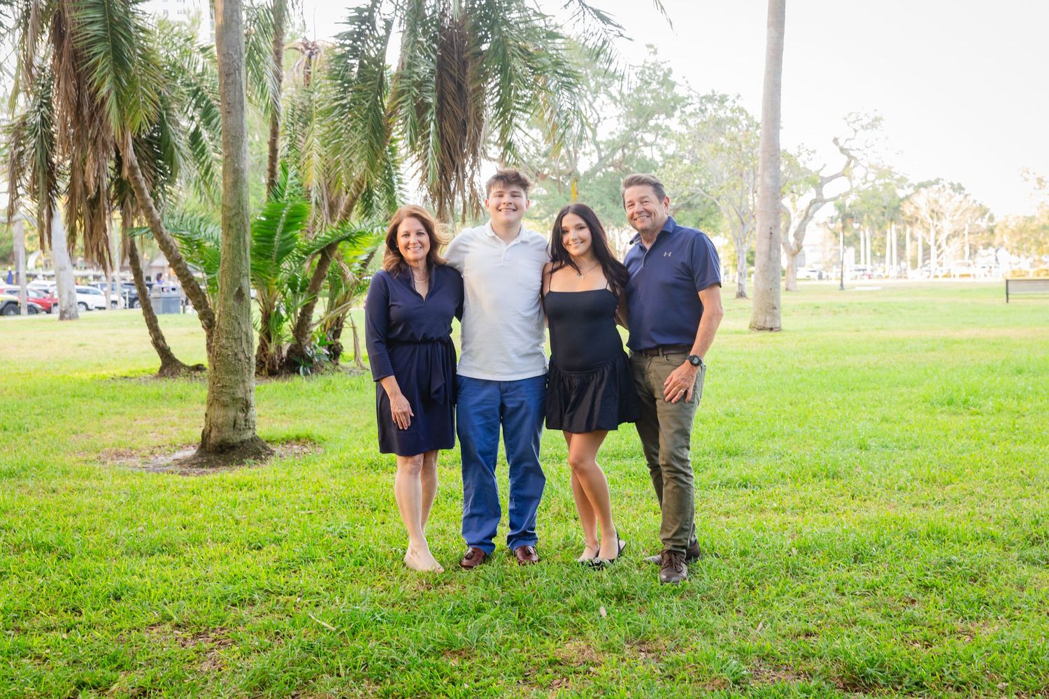 Family of four smiling together outdoors in a lush green park with palm trees on a sunny day.