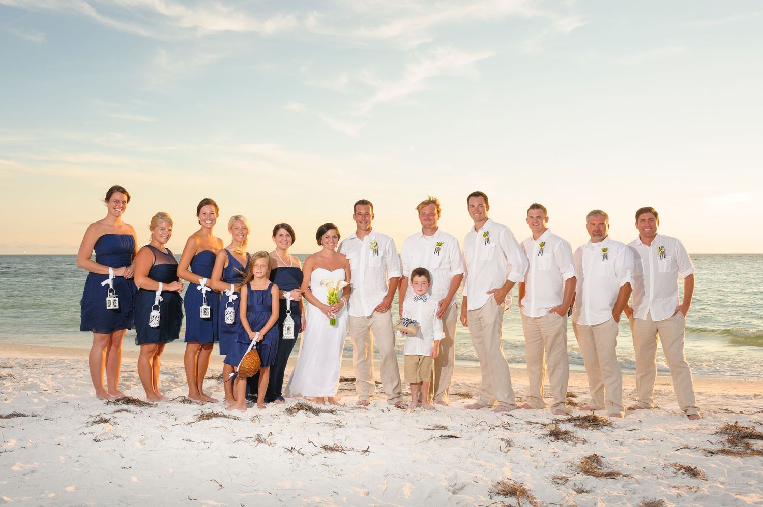 Wedding party poses on a sunny beach, bridesmaids in navy blue dresses and groomsmen in white shirts with tan pants.