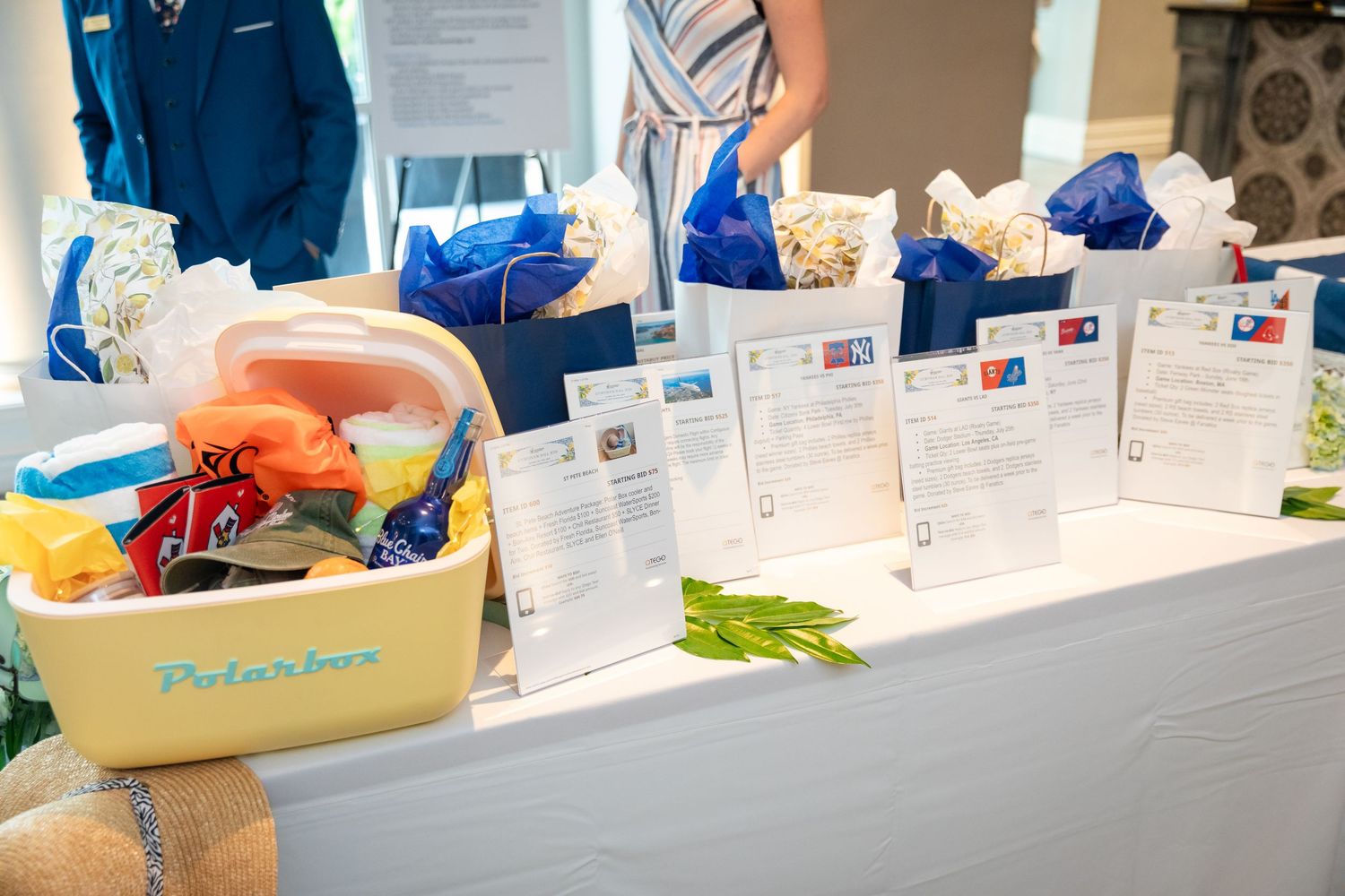 Silent auction table displaying gift baskets and blue bags with descriptions at an indoor charity fundraising event.