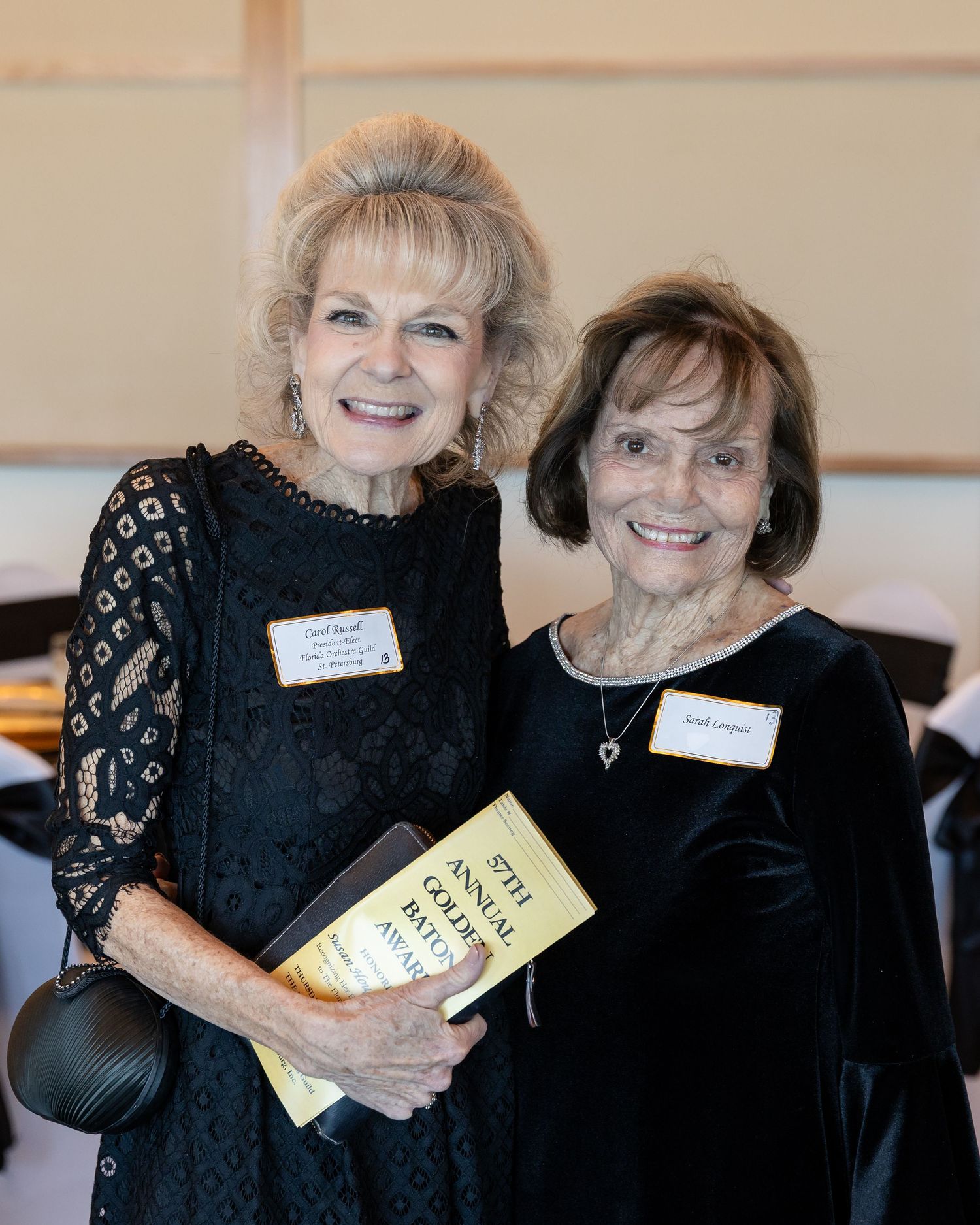 Two elegantly dressed women in black attire smile together at an annual golf awards banquet event.