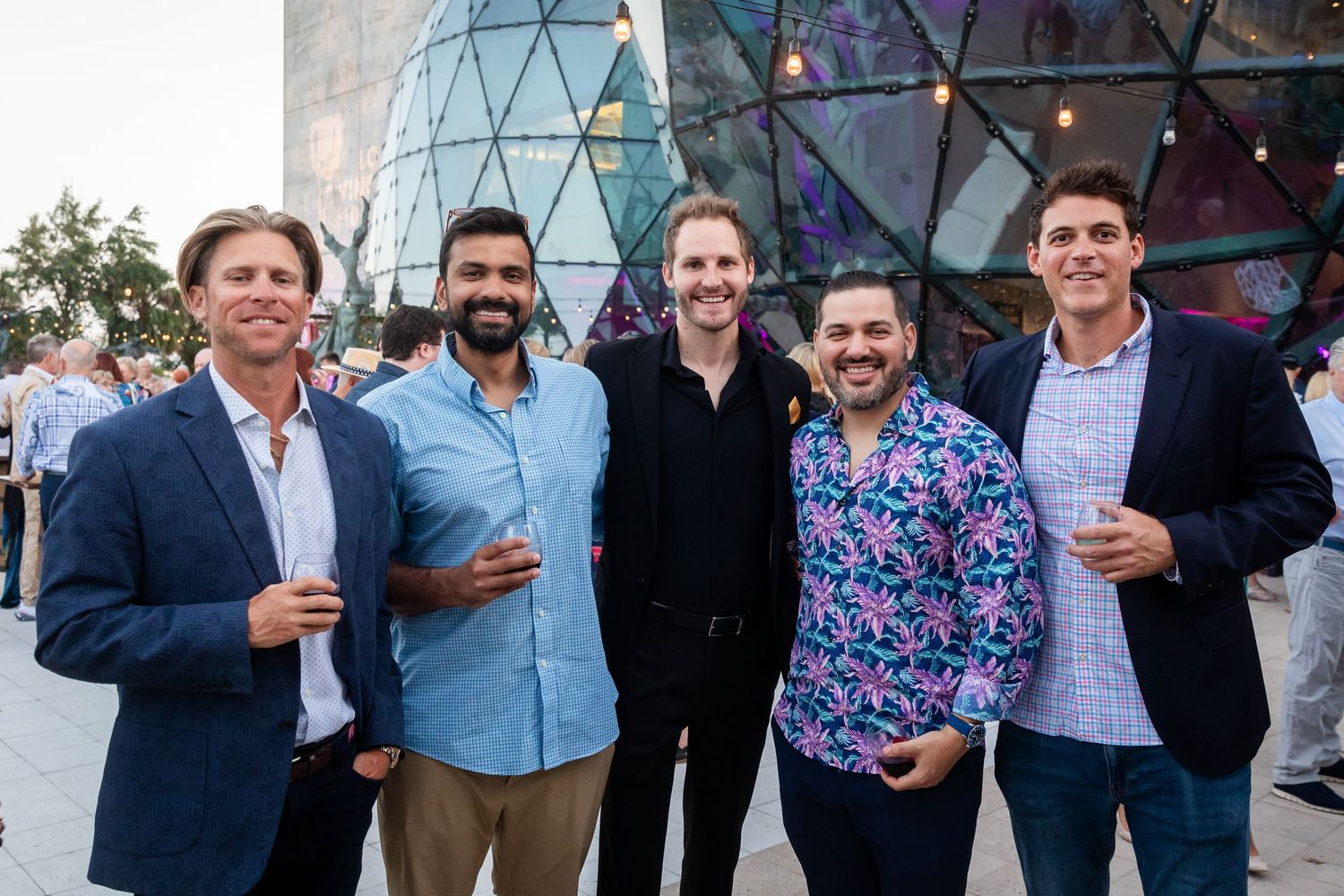 Five men smiling and holding drinks at an outdoor evening event near a modern geometric glass building.