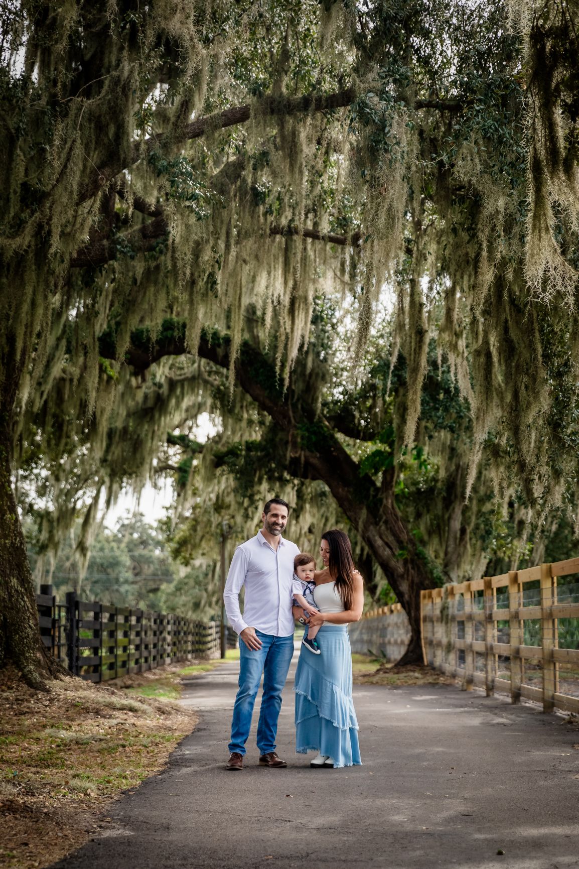 Family of three poses on a tree-lined path draped with Spanish moss, with a wooden fence and lush greenery in background.