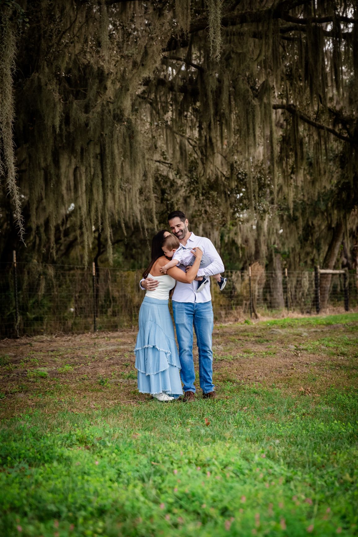 Couple embracing outdoors under Spanish moss-draped trees, woman in blue dress and man in white shirt and jeans.