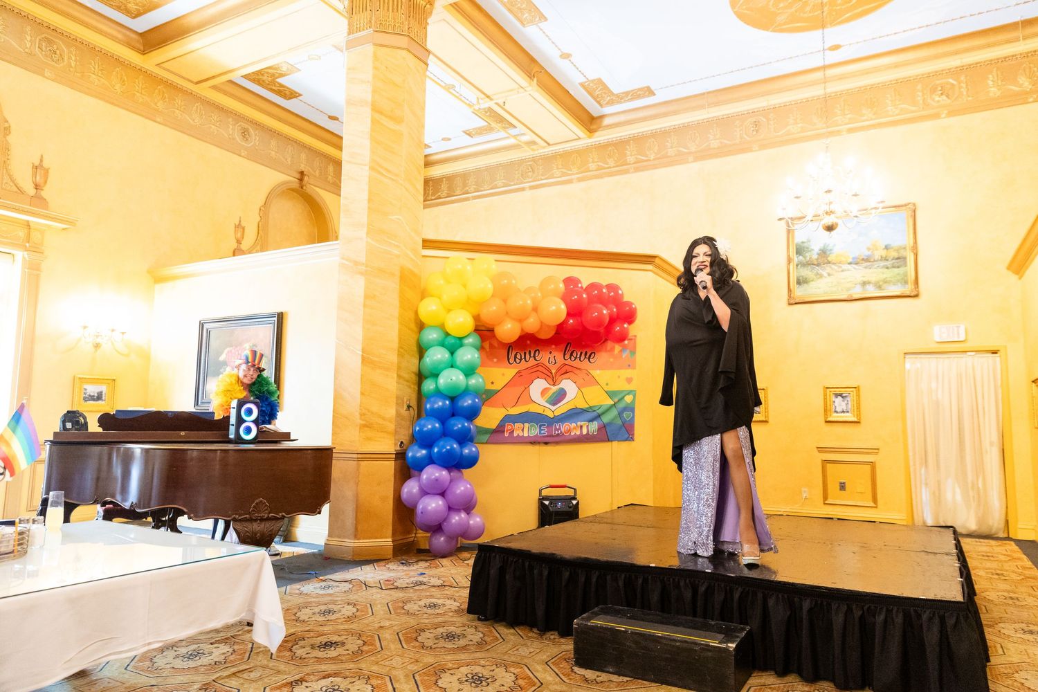 Drag performer on stage with rainbow balloon arch backdrop in an elegant gold ballroom venue.