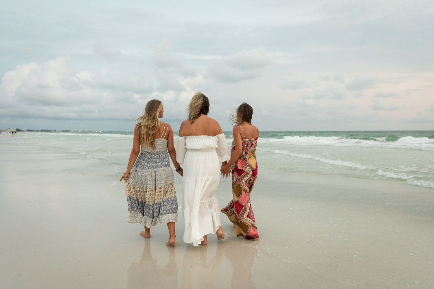 Three women in flowing dresses walk along a sandy beach toward ocean waves under a cloudy sky.