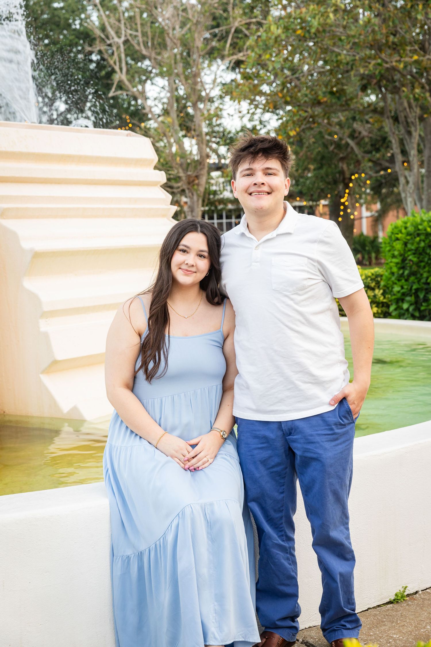 A young couple smiling in front of a fountain, woman in blue dress and man in white polo shirt and jeans.