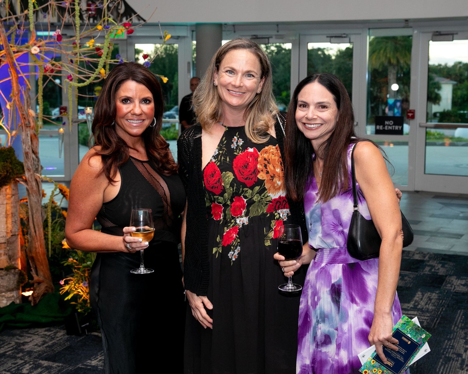 Three women smiling and holding wine glasses at an elegant evening gala event.