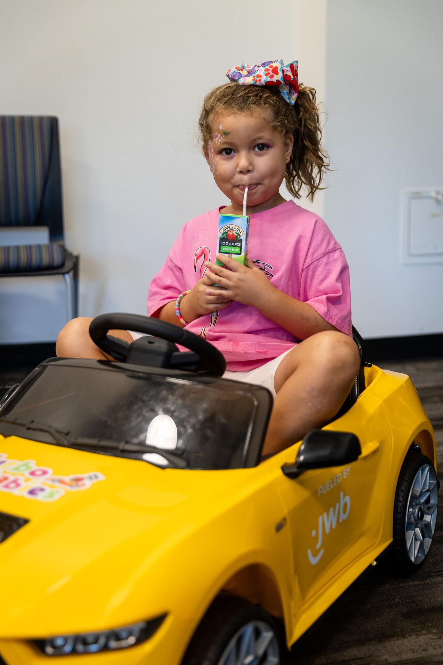 Young girl in pink shirt sips juice box while sitting in yellow JWB toy electric car with bow in hair.