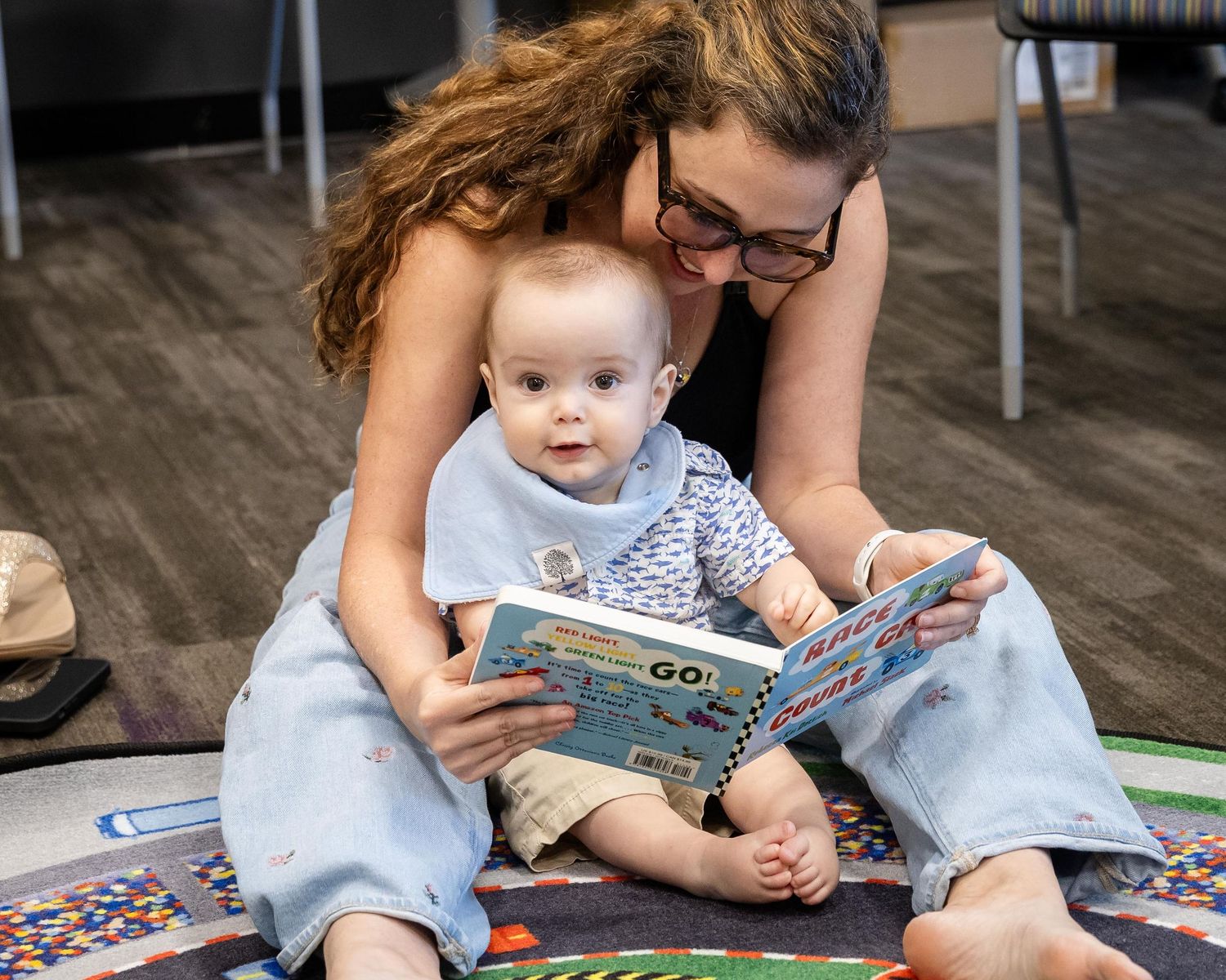 A mother sits on a colorful rug reading a picture book with her curious baby on her lap.
