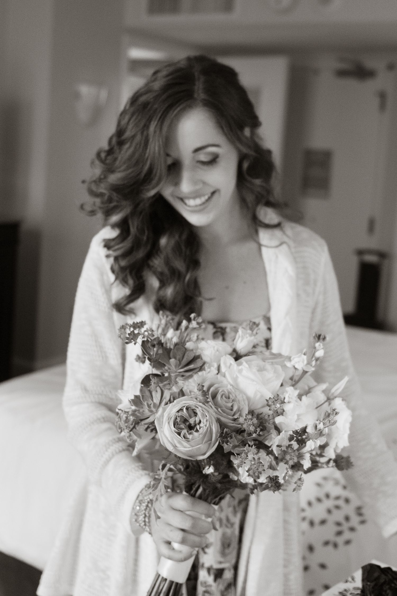 Black and white photo of a smiling bride in a robe holding a beautiful floral bouquet while getting ready.