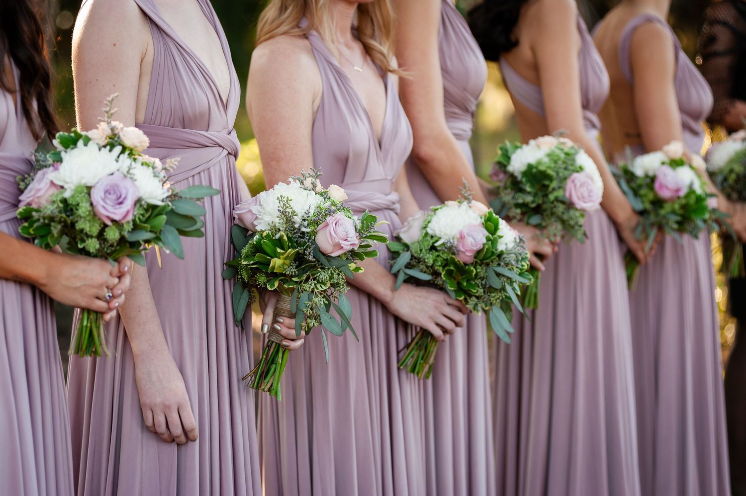 Row of bridesmaids in dusty lavender wrap dresses holding elegant white and pink rose bouquets with greenery.