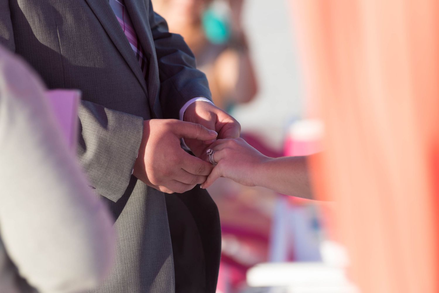 Close-up of hands clasped together during an outdoor wedding ceremony with colorful decorations in background.