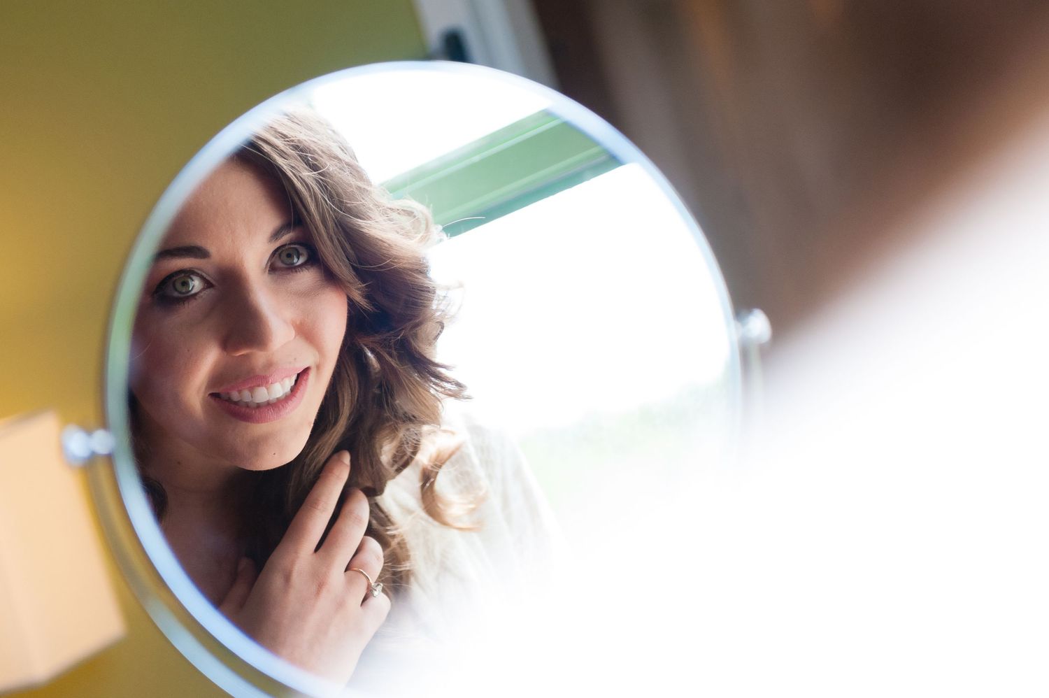 Bride admiring her curly hair and makeup in a round mirror while getting ready for her wedding day.
