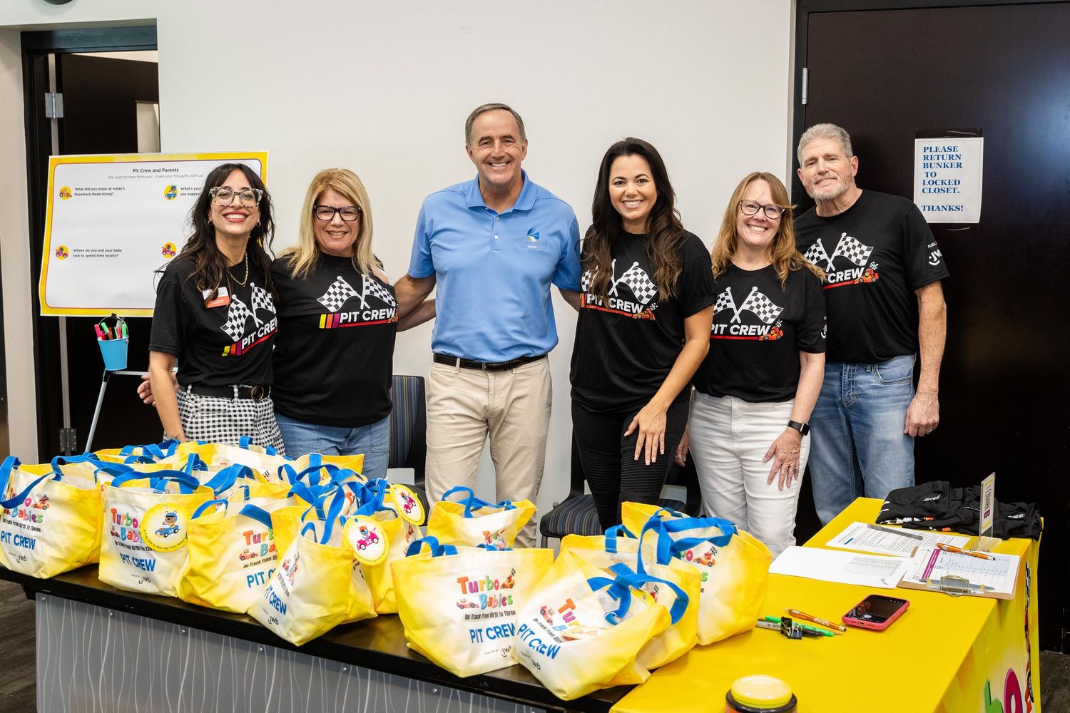 Group of six volunteers in matching black shirts posing with donated food bags at a community charity event indoors.