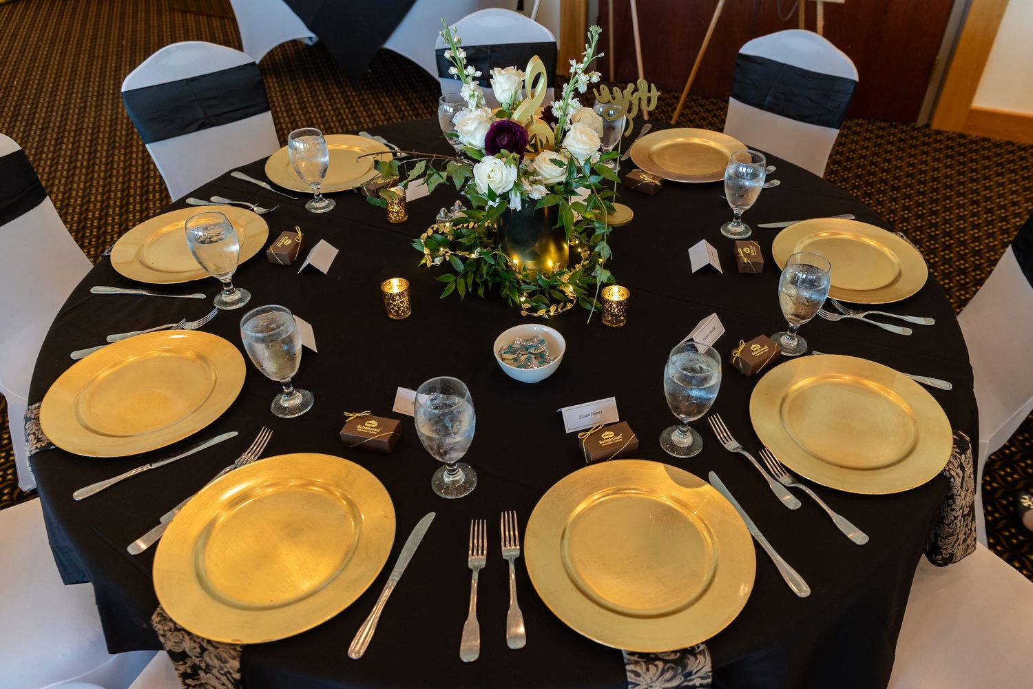 Elegant round banquet table with gold charger plates, black tablecloth, floral centerpiece, and silver cutlery for a formal event.