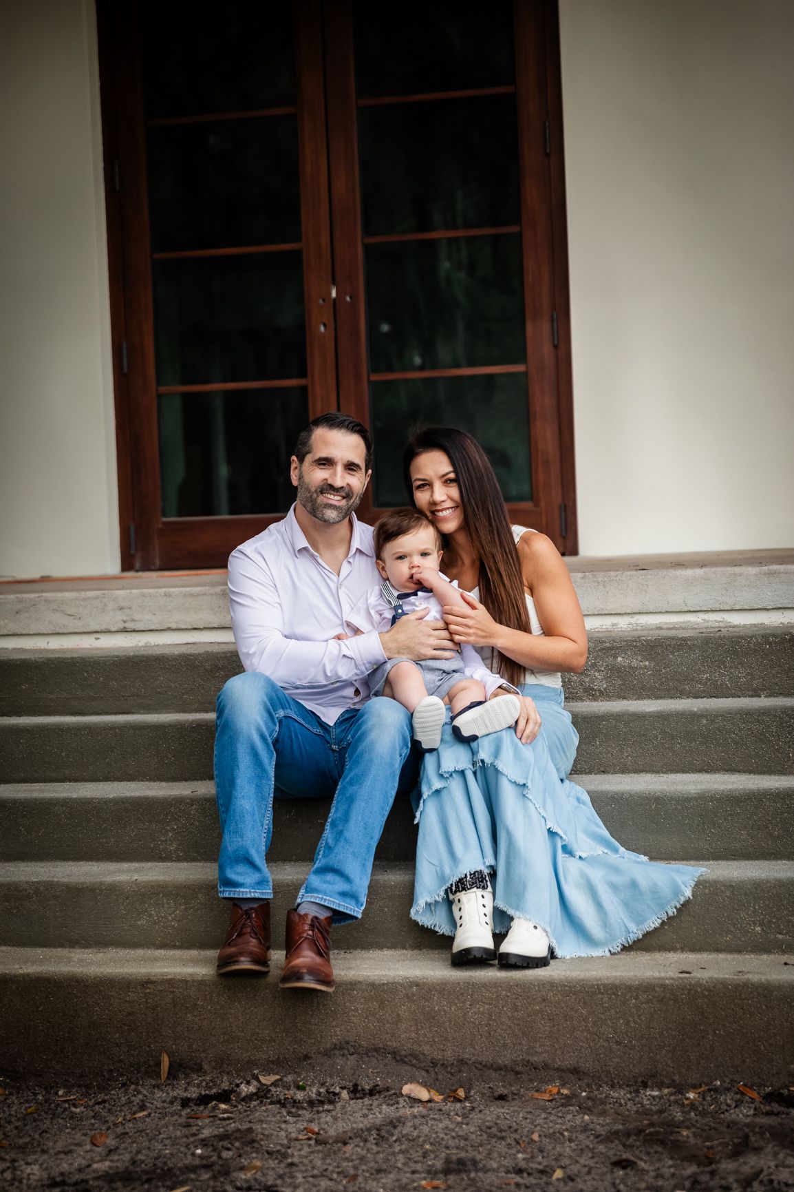 A smiling family of three sits on stone steps, parents holding their baby boy in front of a building entrance.