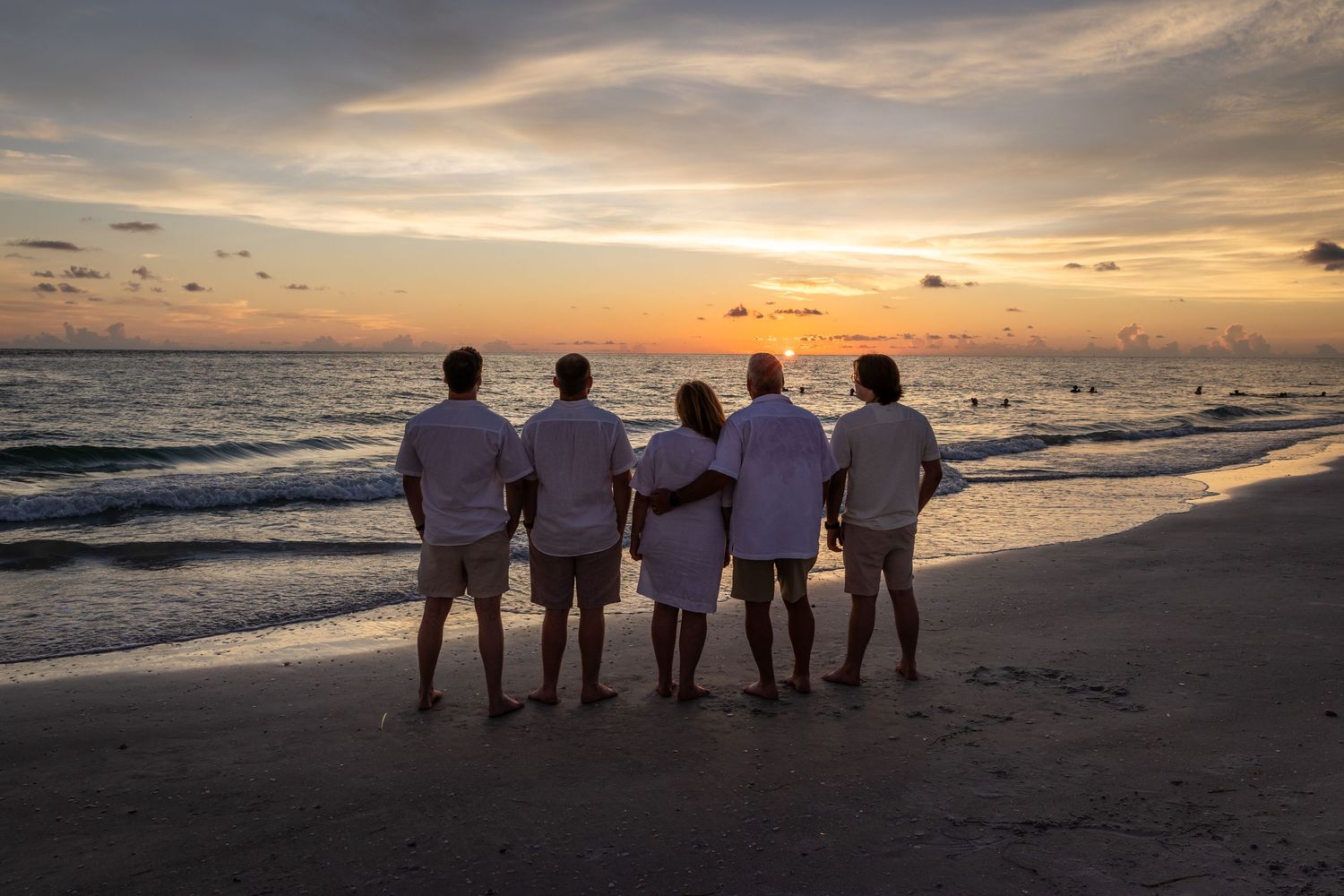 Family of five in white outfits watching a stunning sunset on a sandy beach with colorful skies and calm ocean waves.