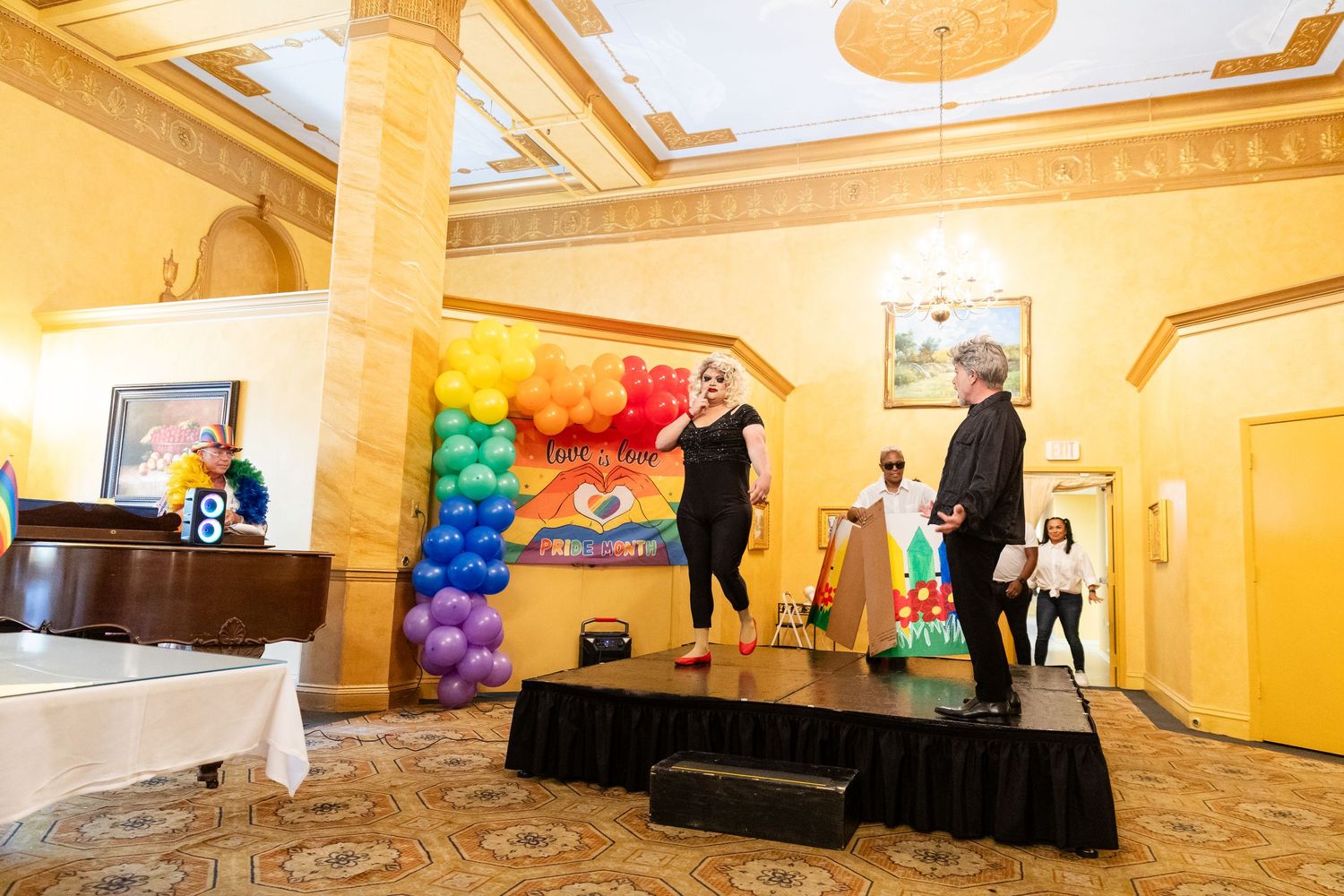 Performer on stage with rainbow balloon arch backdrop at a Pride-themed indoor celebration event.