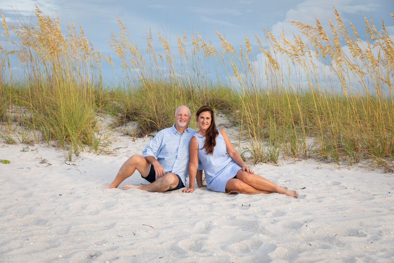 Couple sitting together on white sand beach dunes with sea oats, smiling in casual blue outfits on a sunny day.