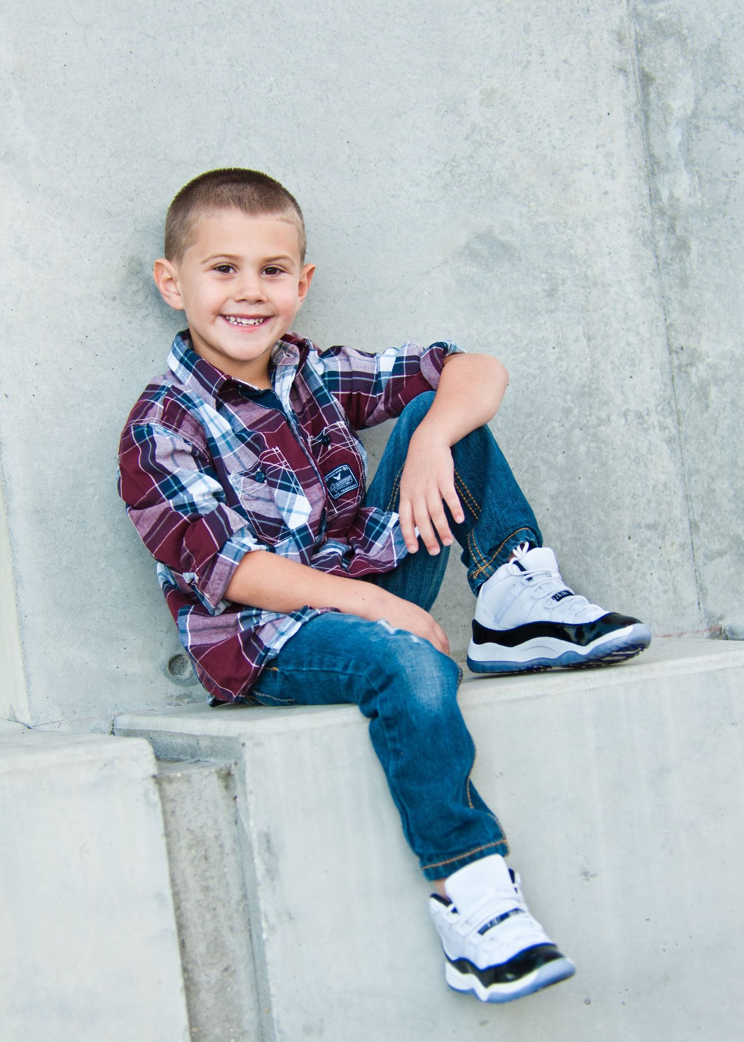 Smiling young boy in plaid shirt and jeans sitting on concrete steps wearing Air Jordan sneakers.