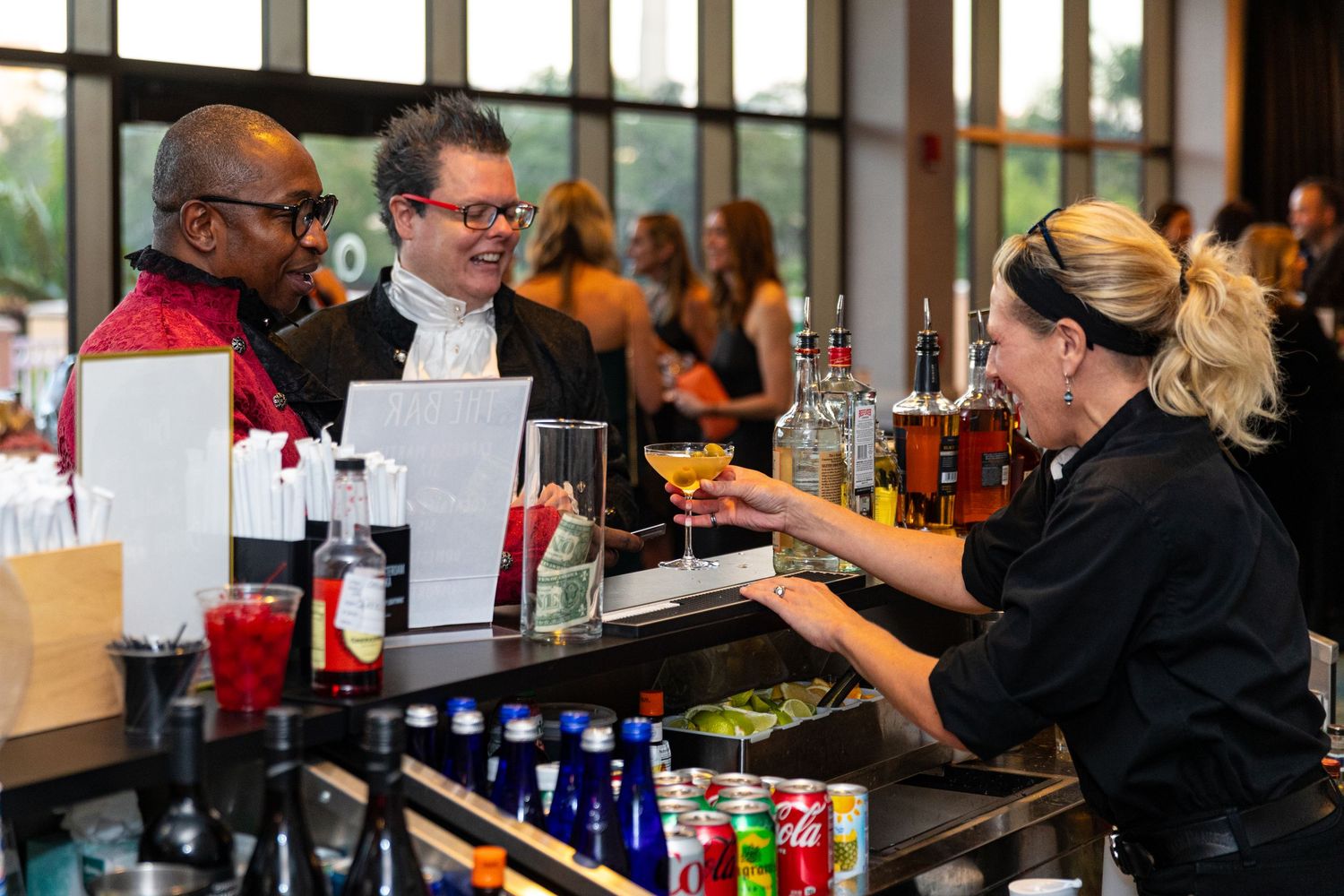 Bartender serving cocktails to two smiling guests at a well-stocked bar during a lively event.